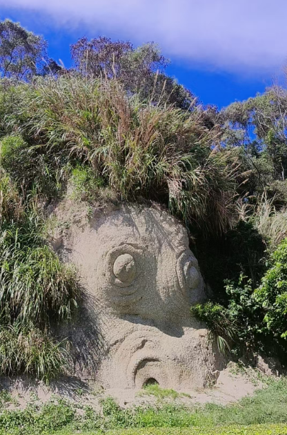 A large stone sculpture of a fish with prominent eyes and mouth, surrounded by lush greenery and wild grass under a bright blue sky, matches the post's description of a "鲤鱼石" (Koi Fish Stone) photographed at Pingtan Island, symbolizing prosperity and good fortune ("见鱼发财") for the start of the work week.