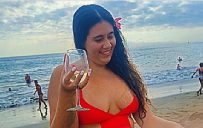 A woman in a red bikini on a beach holding a glass of white wine, smiling at the camera with the ocean and other people visible in the background.