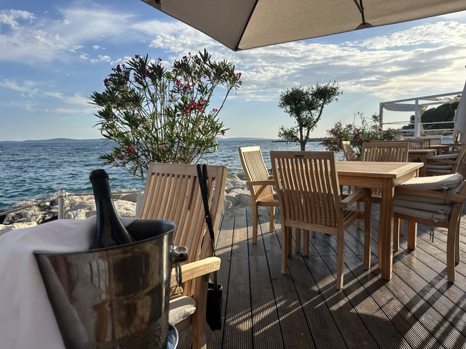 The image shows a wooden deck with several chairs and tables arranged around it. There are two umbrellas providing shade over some of the seating areas. A bottle of wine can be seen next to one of the chairs, suggesting that this could be an outdoor dining area or a patio setting for enjoying a meal by the water.

The post about spending Zcash in Split, Croatia is related to the image because it features a restaurant with wooden furniture and a view overlooking the ocean. The combination of the wine bottle and the outdoor seating arrangement creates a relaxing atmosphere that would be perfect for someone looking to unwind and enjoy some good food and drink while taking in the beautiful scenery.