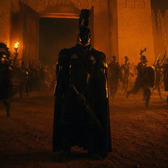 A dark, armored figure with a plumed helmet stands holding a sword in a fiery, smoke-filled courtyard as soldiers run in the background.