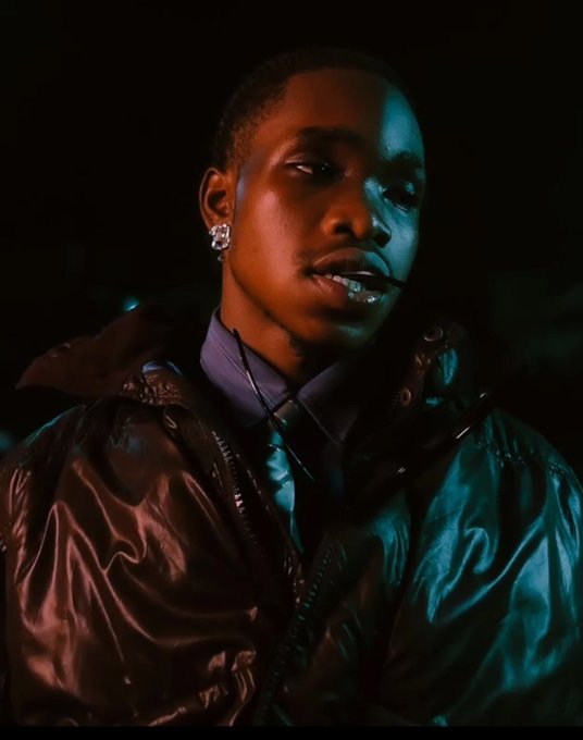 A young man is captured in a stylized portrait, wearing a glossy dark puffer jacket and an earring, with dramatic teal and warm lighting illuminating his face against a black background.