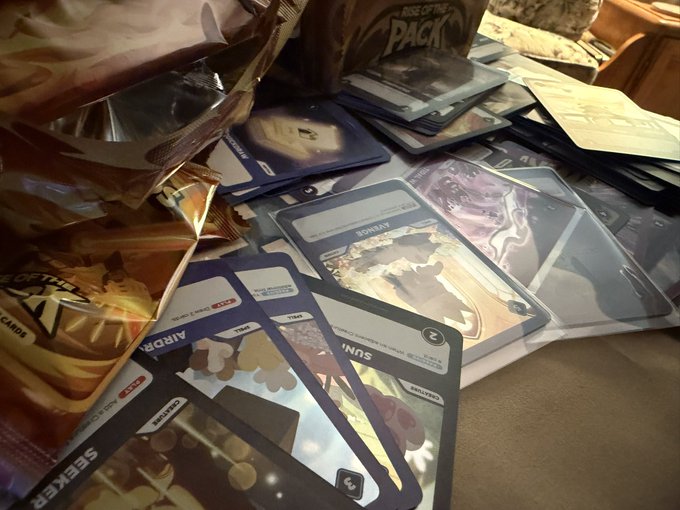A collection of trading cards from 'Rise of the Pack' is scattered on a surface, with booster packs and foil-wrapped cards visible, including cards labeled 'Ardyn' and 'Seeker'.