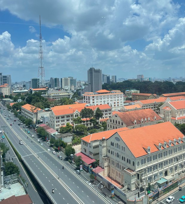 An aerial cityscape shows a wide avenue lined with French colonial-style buildings featuring distinctive orange-tiled roofs, set against a backdrop of modern skyscrapers and a tall communication tower under a partly cloudy sky. The vibrant, sunlit view captures the energetic urban charm that complements the post's hopeful Monday greeting wishing for a productive and "green" week ahead.