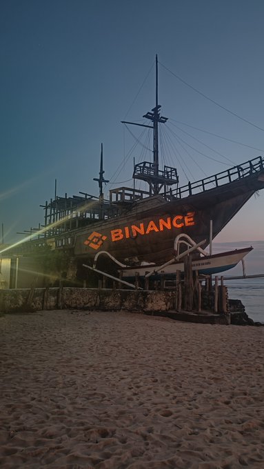 A pirate ship sits on a sandy beach at dusk with the word "BINANCE" illuminated in orange on its hull, likely referencing the crypto exchange Binance.