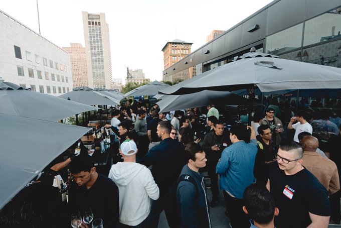 A crowded outdoor rooftop bar scene with numerous patrons gathered under gray umbrellas, set against a city skyline backdrop.