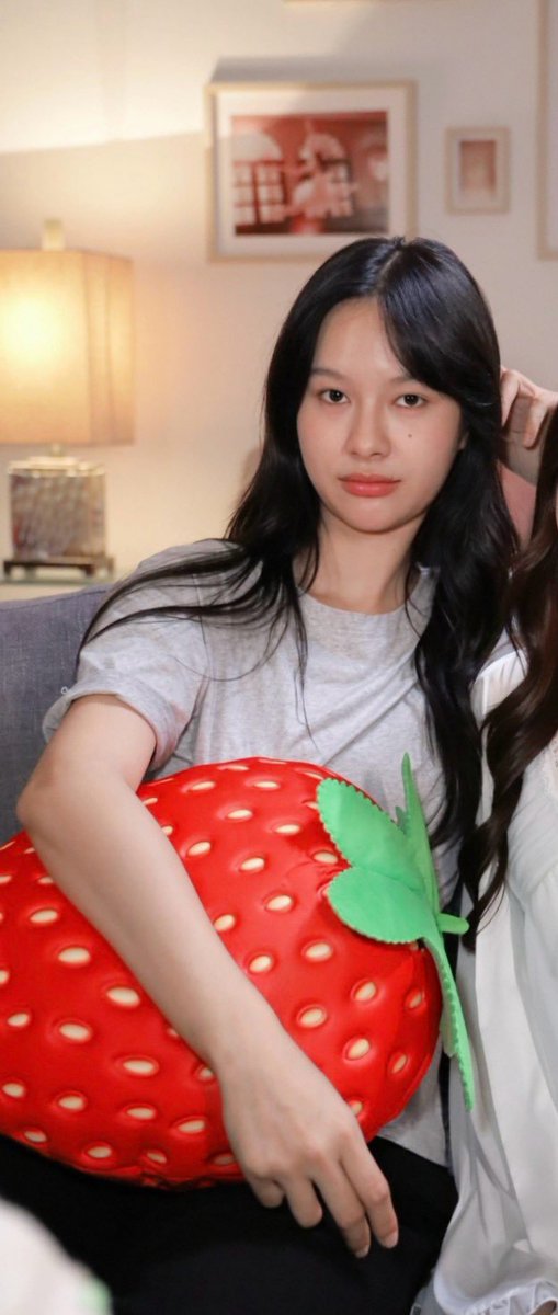 A young woman with long dark hair sits on a couch holding a large plush strawberry pillow, while another person in white touches her hair. The post appears to reference a Thai drama series "DITTO" and playfully discusses character archetypes and makeup choices, with the strawberry pillow possibly symbolizing a "strawberry girl" aesthetic.