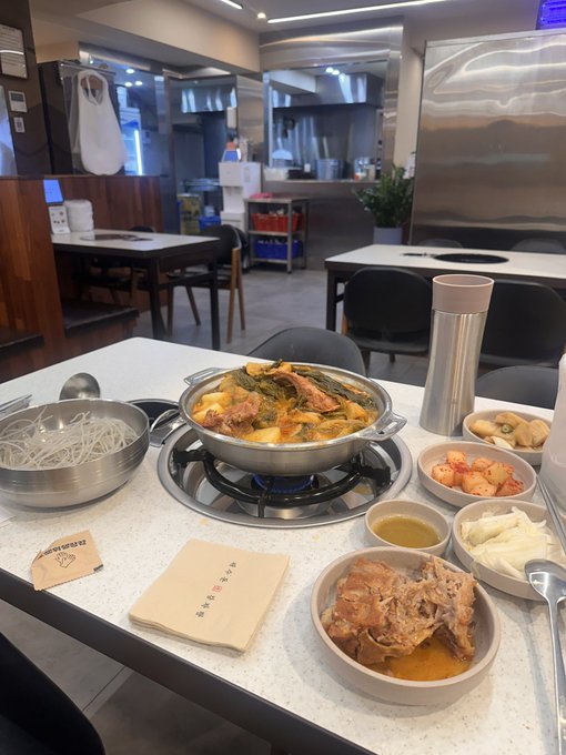 A restaurant table set with a steaming Korean hot pot, side dishes of bean sprouts, kimchi, radish, and dipping oil, along with a thermos and napkins.