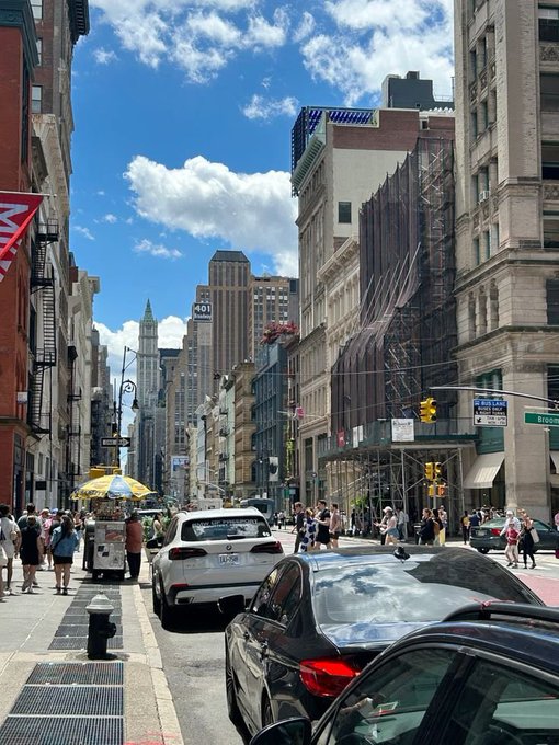 The image shows a busy city street with tall buildings lining both sides, including one building that has scaffolding covering its windows. The scene captures the hustle and bustle of urban life as people walk along the sidewalks or wait at bus stops. There are several cars parked on the side of the road, adding to the sense of activity in the area. A person is seen walking near a car with a "Good Afternoon" sign attached to it, indicating that the time captured in the image might be around mid-day. The post also mentions that the 25 quacks era is over for those who have been receiving low quacks on @wallchain these days.
