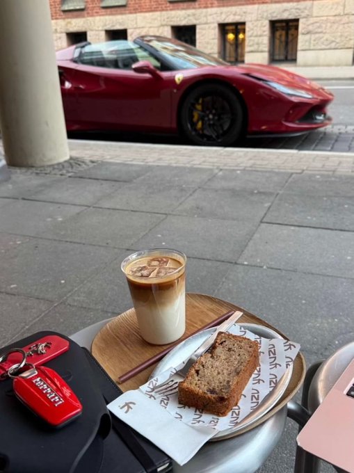An outdoor cafe scene featuring an iced coffee, a slice of cake, and a car key fob on a round table, with a red Ferrari parked on the street in the background.