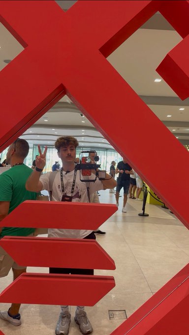 A young man with curly hair flashes a peace sign while holding up a phone to take a mirror selfie, framed by a large red sculptural structure in what appears to be an airport or mall.
