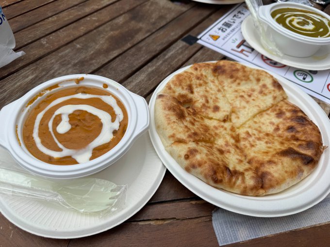 A plate of cheese naan sits beside a bowl of chicken curry garnished with a swirl of cream, served on a wooden table alongside a cup of green soup. The Japanese menu and place settings suggest a Japanese restaurant, matching the post about celebrating Bitcoin lending rewards with a curry and naan meal.