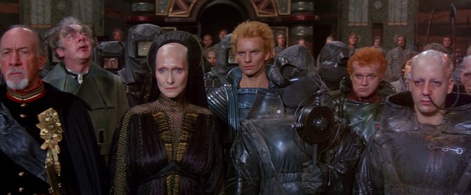 A scene from the movie Dune featuring characters including Baron Harkonnen, Princess Irulan, Feyd-Rautha, and Glossu Rabban standing together in elaborate costumes.
