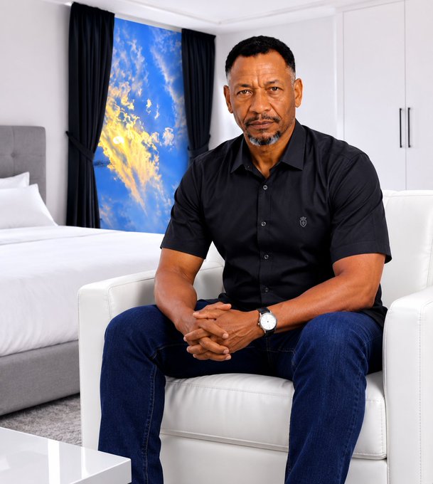 A confident man in a black shirt and jeans sits with hands clasped in a modern, bright bedroom setting. The "A New Day!" post pairs with his poised, forward-looking expression, suggesting readiness to pursue ambitious goals head-on.