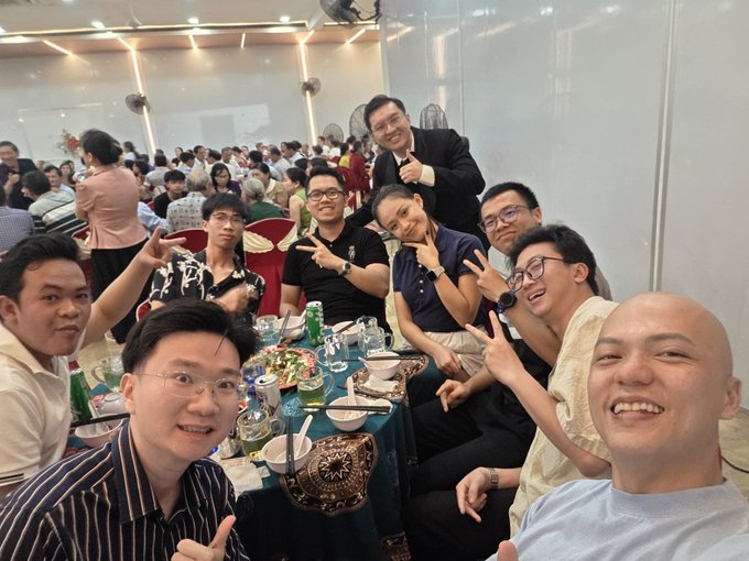 A group photo of around 10 men smiling and posing with peace signs at what appears to be a restaurant gathering or banquet, with food and drinks on the table.