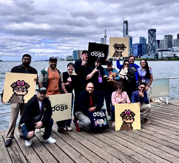 A diverse group of people pose on a wooden pier by a waterfront city skyline, holding pixelated dog artwork and "dogs" branded signs. The image relates to the post by capturing the camaraderie of the Doginal Dogs community gathering at the Blockchain Futurist event, where members proudly display their NFT artwork to celebrate their shared connection.