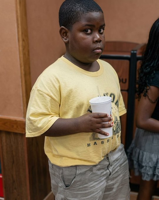 A young boy in a yellow t-shirt with graphics holding a white styrofoam cup, standing indoors near another person in a gray skirt.