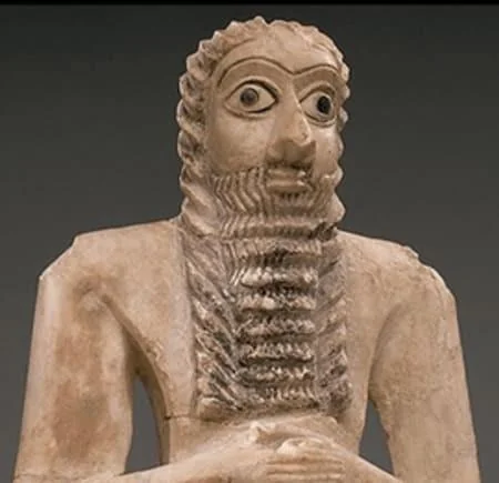 An ancient, light-colored stone statue of a bearded figure with large, prominent eyes and textured clothing, displayed against a dark background.