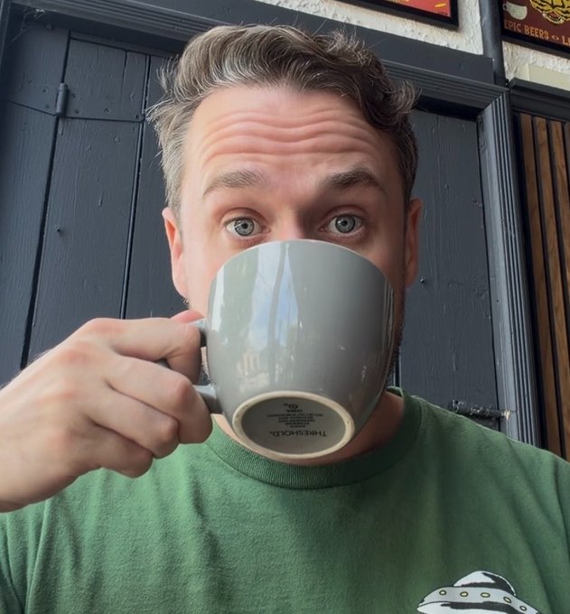 A man with light hair drinks from a gray "miHArGeBACK" mug while wearing a green shirt in front of dark doors.