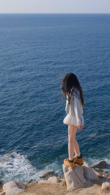 A young woman with long dark hair, wearing a white top, shorts, and brown boots, standing on rocks overlooking the blue ocean.