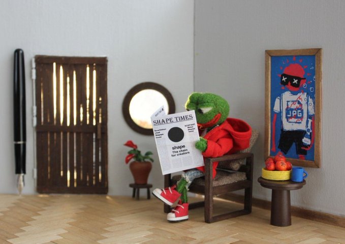 A green felted frog character in a red hoodie and red sneakers sits in a miniature wooden chair reading a newspaper titled "SHAPE TIMES" with the headline "shape: the chain for creators," while a framed poster on the wall displays a character wearing a shirt labeled "JPG." The whimsical diorama creatively plays on the "GM" (good morning) crypto greeting and the concept of "CC0" (Creative Commons Zero) public domain content, with the "SHAPE TIMES" newspaper and "JPG" shirt symbolizing the free, creator-focused ethos of CC0 culture.