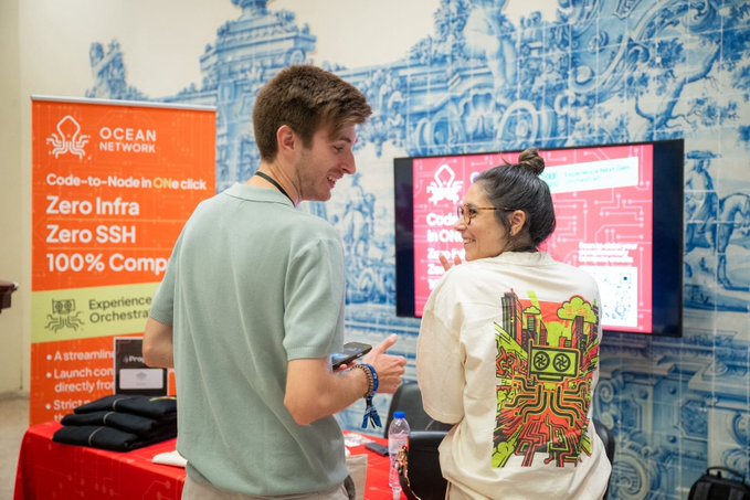 Two people chatting at an Ocean Network promotional booth, with a banner advertising features like 'Code-to-Node in One click', 'Zero Infra', 'Zero SSH', and '100% Compliance', set against a blue and white patterned wall.