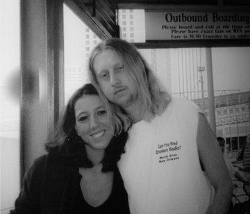 A black and white photo shows a smiling couple posing together in front of a sign that reads "Outbound Boarding" and "Please have exact fare of $3.75," with the woman wearing a T-shirt that says "Can You Find Damian Kulash," accompanying a post celebrating a couple's 23rd anniversary and reflecting on their long-lasting relationship.