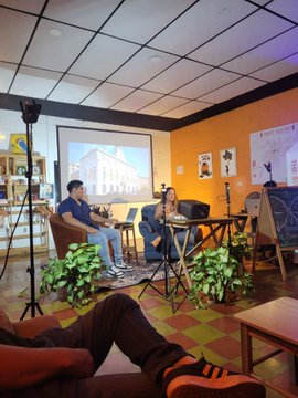 Two people sit in chairs on a stage in a casual interview setting, with one playing a guitar while a projection screen and audience member's legs are visible in the foreground.