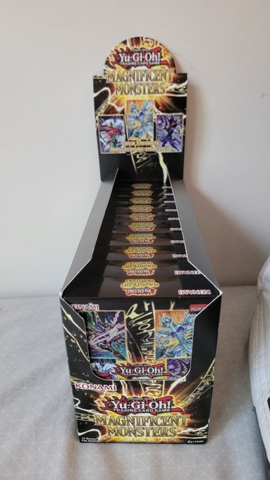 A sealed Yu-Gi-Oh! Magnificent Monsters booster box sits on display, with Konami branding visible on the side and front. The caption reflects the meme of rotating meme coin profits into tangible cardboard assets too nice to open.