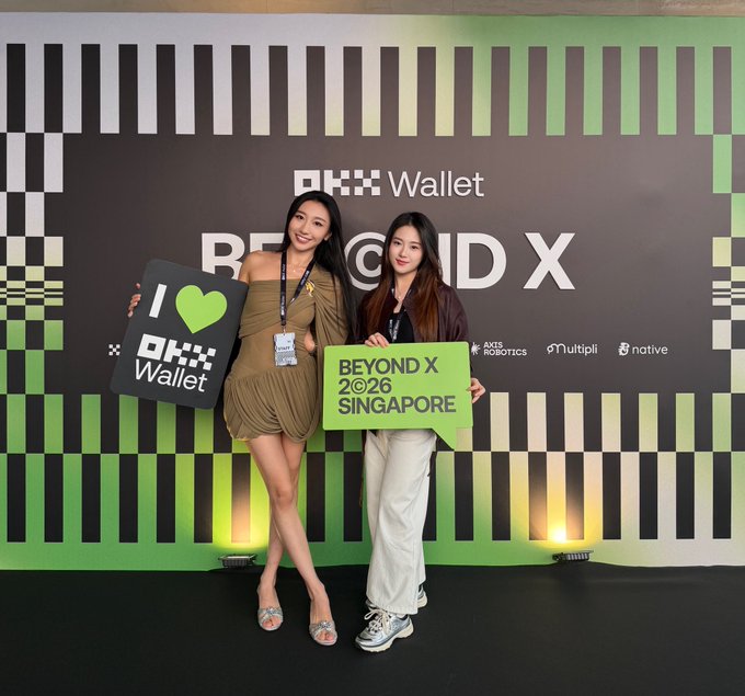 Two women posing at the OKX Wallet 'BEYOND X 2026 SINGAPORE' event, holding a black 'I ❤ OKX Wallet' sign and a green 'BEYOND X 2026 SINGAPORE' sign in front of a striped backdrop.