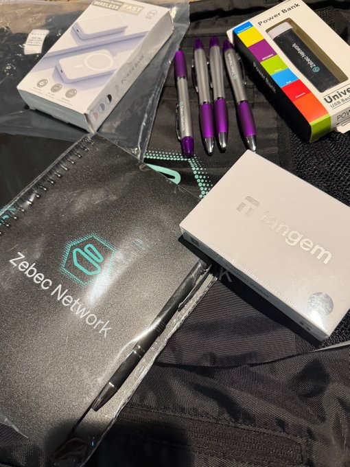 A collection of giveaway items laid out on a black surface, including a "Zebec Network" branded bag, a "Tangem" wallet box, four purple and silver pens, a wireless charger box, and a "Power Bank" box — matching the Zebec_HQ-themed giveaway the builder teased.
