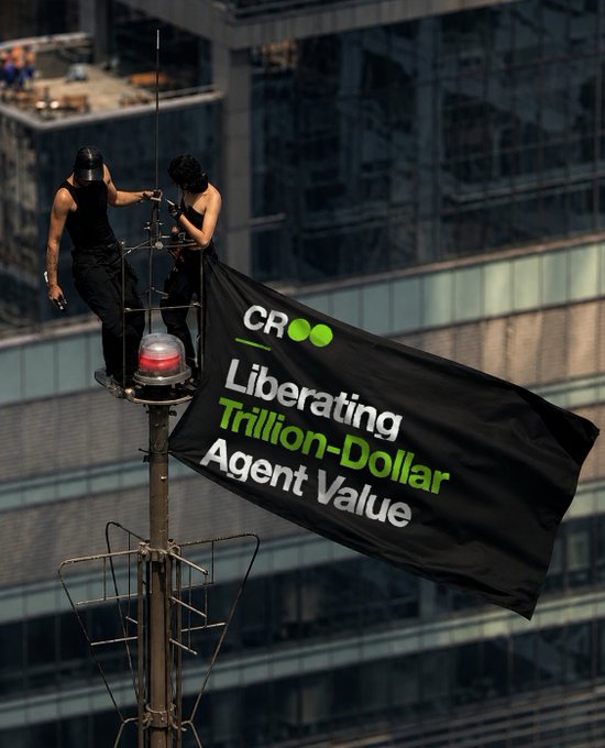 Two activists dressed in black and masks scale a tall pole or antenna atop a city building, unfurling a large black banner that reads "CRoo Liberating Trillion-Dollar Agent Value," a bold protest-style statement echoing the post's affirmation of "True." The dramatic high-rise setting emphasizes the activist visual rhetoric, framing the message as a defiant declaration about unlocking trillion-dollar value in the agent economy.