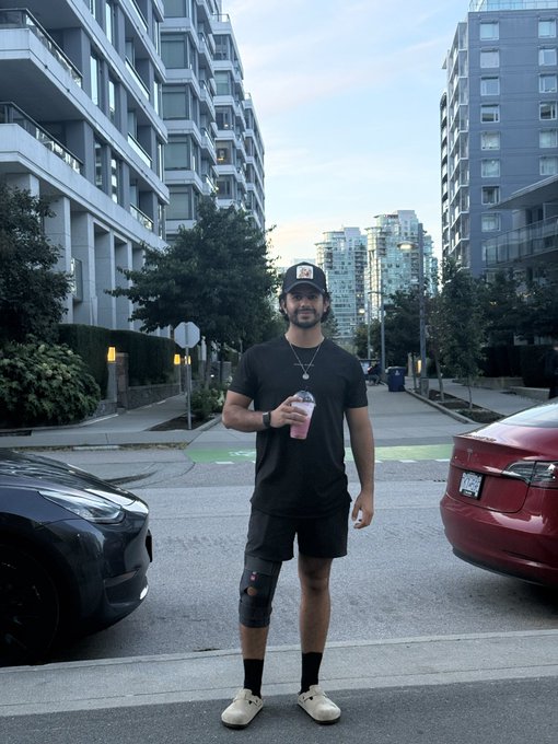 A man in a black cap, dark t-shirt, and shorts stands on an urban street holding a pink drink, with two cars flanking him and high-rise buildings in the background. The post credits @serdre_, likely the photographer behind the casual streetwear-style portrait.