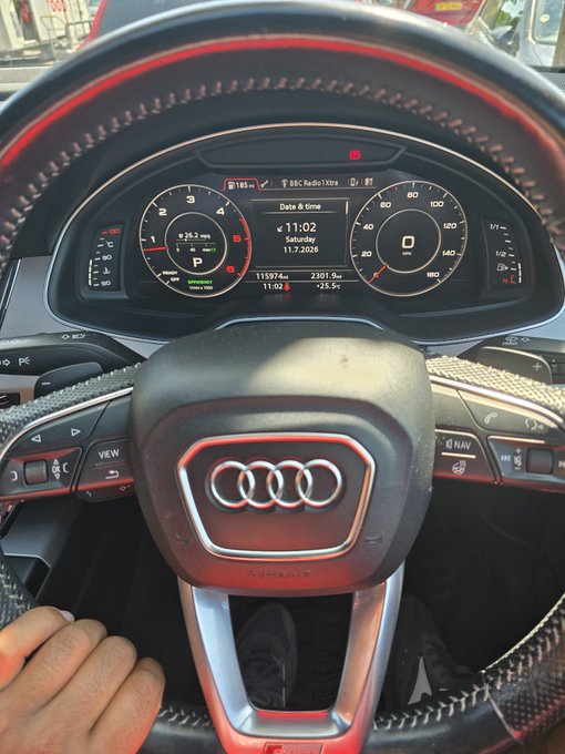 A view of an Audi steering wheel with the digital dashboard showing 'Date & time 11:02', 'Saturday 11.7.2026', a speed of 0, and other vehicle metrics including '115974 km' and '23019 km'.