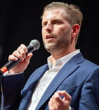 A man in a blue suit jacket and white shirt speaking into a microphone while gesturing with his other hand.