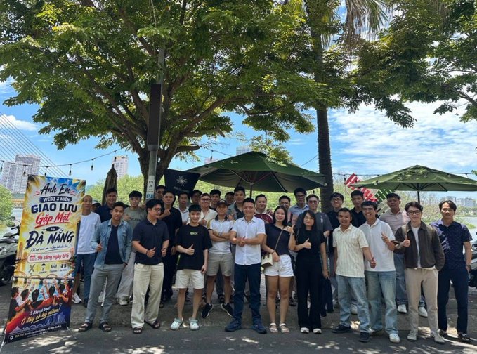 The image features a group of people posing for a photo, standing under an umbrella near trees. The group consists of men and women wearing casual clothes, including white shirts and black pants or skirts. They are gathered together with smiles on their faces, indicating that they might be friends or acquaintances. In the background, there is a banner promoting an event called "The toughest time is when we unite! Da Nang Web3 Meetup is a casual but insightful event where people from different backgrounds and histories with the industry share their personal experience". The group appears to be enjoying themselves in this outdoor setting, possibly attending or participating in the web3 meetup.