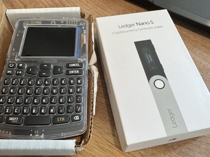A photograph of a homemade DIY hardware wallet next to a commercial "Ledger Nano S Cryptocurrency hardware wallet" box on a wooden desk.
