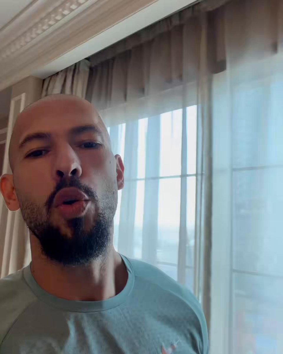 In the image, there is a bald man wearing a blue shirt standing in front of a window with curtains. He has a beard and appears to be making a funny or thoughtful expression as he looks at the camera. The caption above him reads " Andrew Tate predicts that Bitcoin goes down to $26,000." which suggests that the man might be speculating on the future value of Bitcoin based on its current worth in dollars.