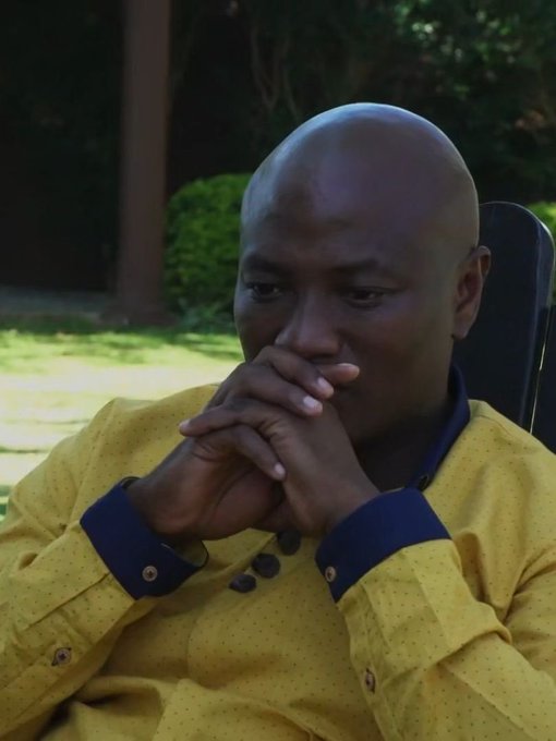 A contemplative man in a yellow dotted shirt with blue cuffs, sitting outdoors with his hands clasped near his face in a prayer-like gesture.