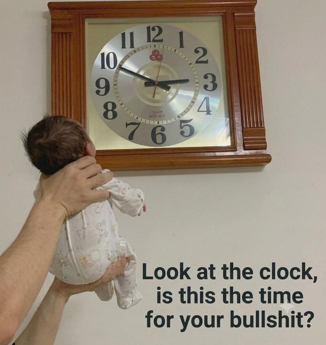 An adult holding a baby up to look at a wall clock, with the humorous caption 'Look at the clock, is this the time for your bullshit?'