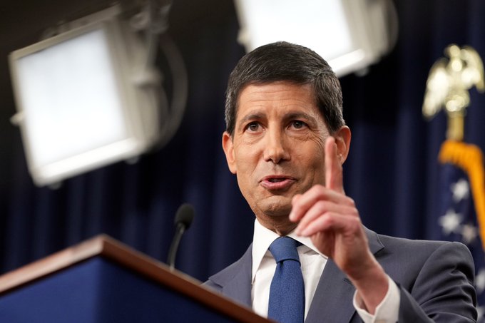 A photograph of a man in a dark suit and blue tie speaking at a podium, gesturing with his index finger raised, with studio lights and an American flag visible in the background.