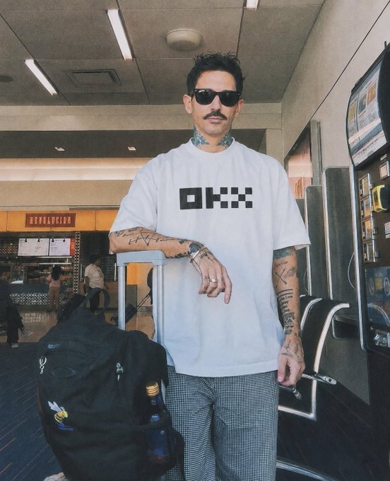 A tattooed man wearing sunglasses and a white t-shirt with a black checkered logo stands in an airport terminal holding a rolling suitcase and backpack.