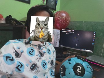 The image features a man sitting at a desk with a computer monitor, keyboard, and mouse. On his shirt, there's a picture of him holding a cat that has been cut out and pasted onto his shirt. The background includes a green wall and a balloon on the wall. There is also an image of a soccer ball in the bottom right corner of the photo.