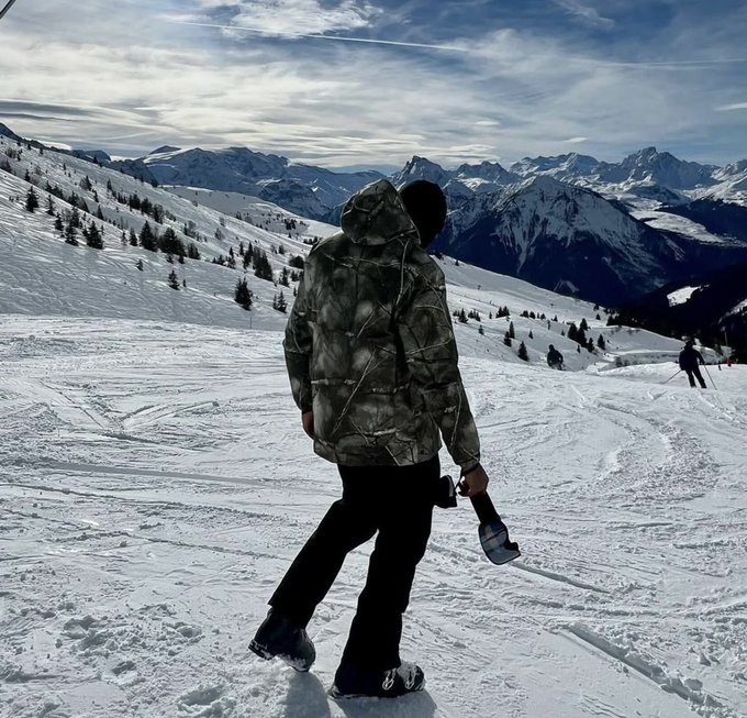 A person wearing a camo jacket and dark pants walks through deep snow on a mountain slope, carrying what appears to be skis or a snowboard, with a vast snowy mountain range in the background.