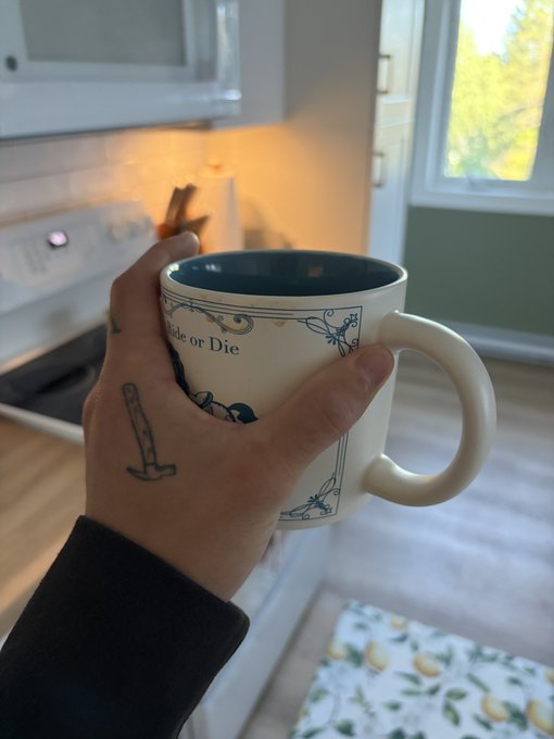 A hand with a sword tattoo on the wrist holds a coffee mug reading 'Ride or Die' in a kitchen setting.