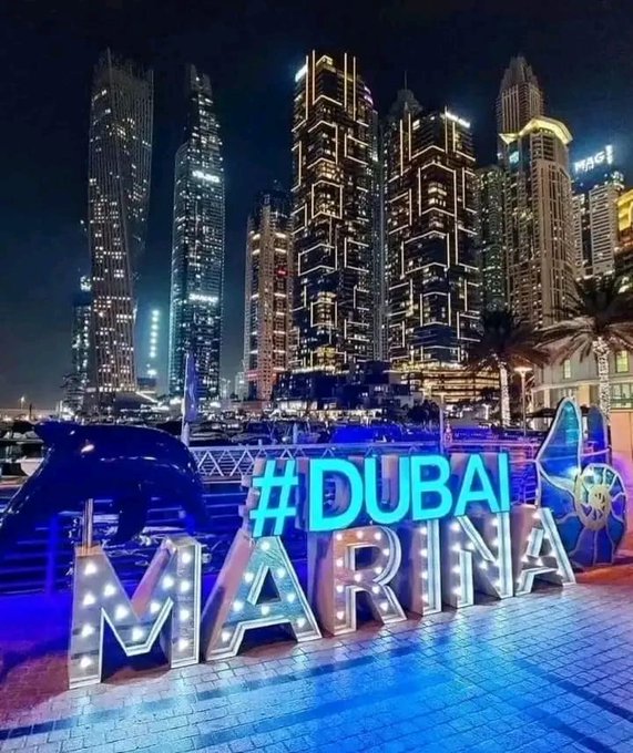 A nighttime view of Dubai Marina featuring tall illuminated skyscrapers and a large decorative '#DUBAI MARINA' sign lit up in blue and white.