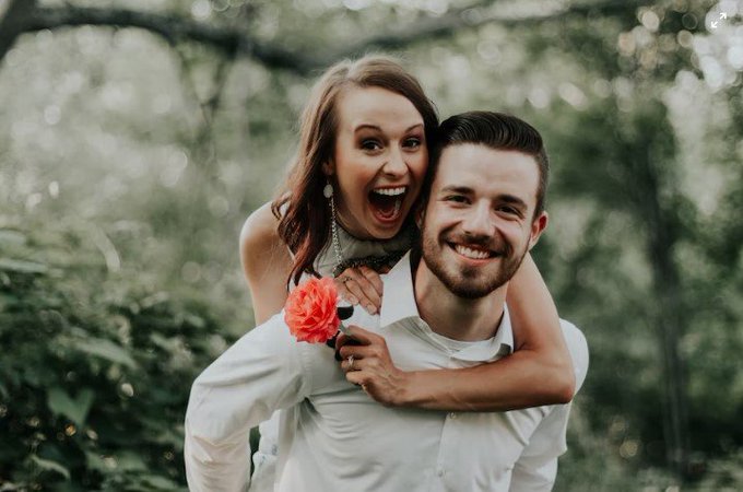 A joyful couple poses for an outdoor engagement photo, with the woman on the man's back laughing excitedly while holding an orange flower, set against a backdrop of green foliage.