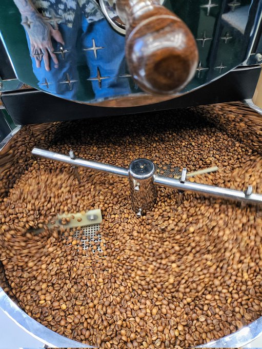 The image shows a large metal machine filled with coffee beans, which appears to be a coffee roaster or grinder. The beans are brown and scattered throughout the machine, indicating that they have been freshly roasted. The scene also includes two people standing near the machine, possibly overseeing its operation or discussing the process of making coffee.