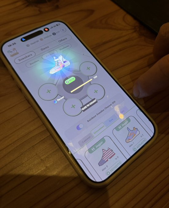 A smartphone on a wooden table displays a sneaker customization app showing different models (Sneakers, Gems, Others), with a 'Rainbow Sneaker Charge: ON' toggle and a 'HOLD TO BOOST' button.