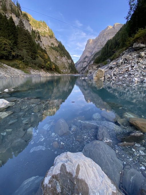 A scenic photograph of a clear alpine lake surrounded by rocky mountains and pine trees, with smooth stones visible beneath the glassy water surface reflecting the blue sky.