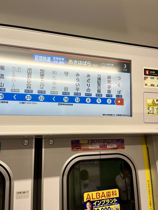 An interior view of a Japanese train with a digital route map showing stations including つくば (Tsukuba) and あきはばら (Akihabara), alongside an ALBA job advertisement poster.